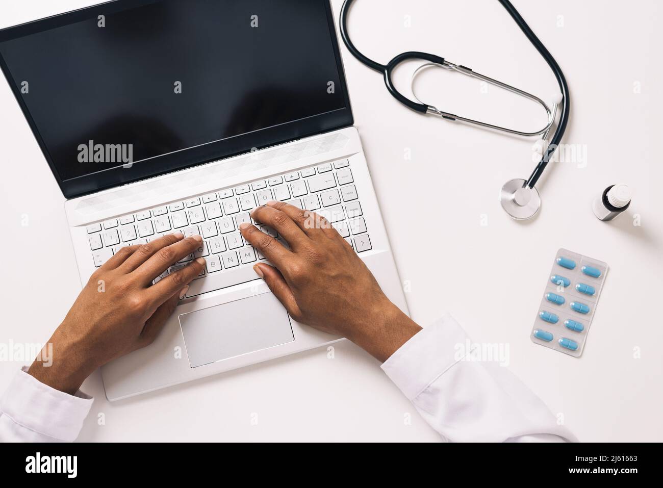 From above crop anonymous African American medical practitioner consulting remotely on netbook at table with stethoscope pills and bottle of syrup Stock Photo