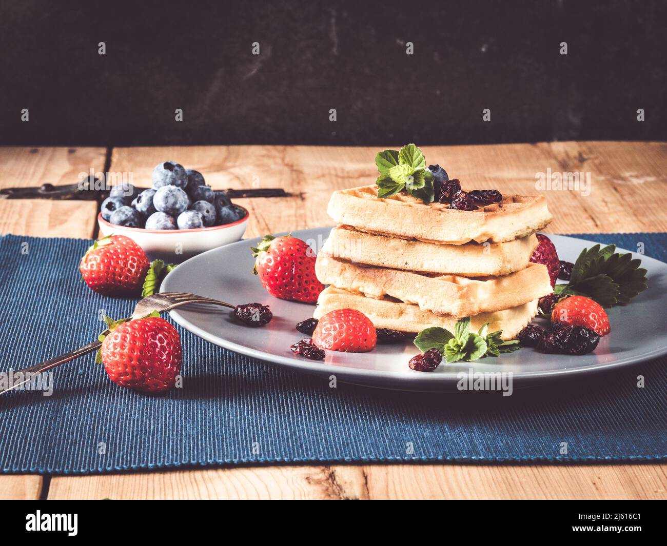homemade waffle stack decorated with blueberries, strwaberries, cranberries and mint; rustic wooden table with placemats and bowls Stock Photo