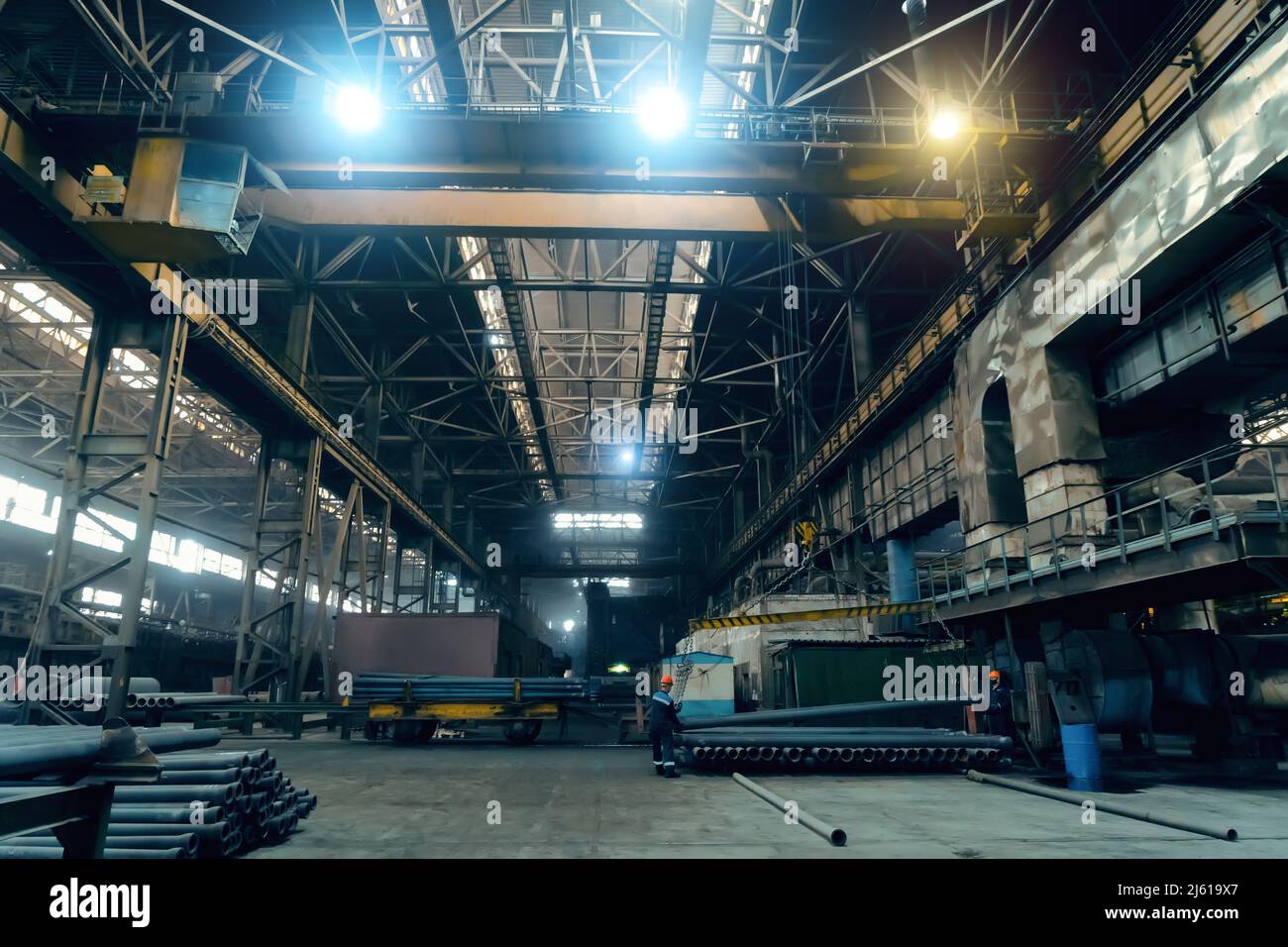 Large girder or beam crane transports iron pipes inside large dark workshop in metallurgical factory. Stock Photo