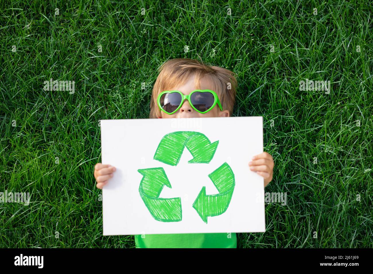 Happy child holding paper with recycle sign outdoor in spring park. Kid lying on green grass. Earth day and ecology concept. Boy holding paper blank Stock Photo