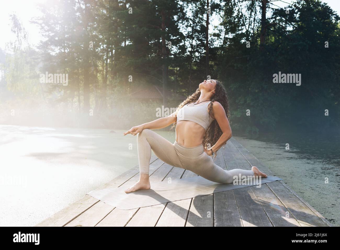 Woman doing Virabhadrasana exercise, warrior pose with knee resting on mat, exercising on a wooden bridge in a park by a pond on a sunny summer mornin Stock Photo