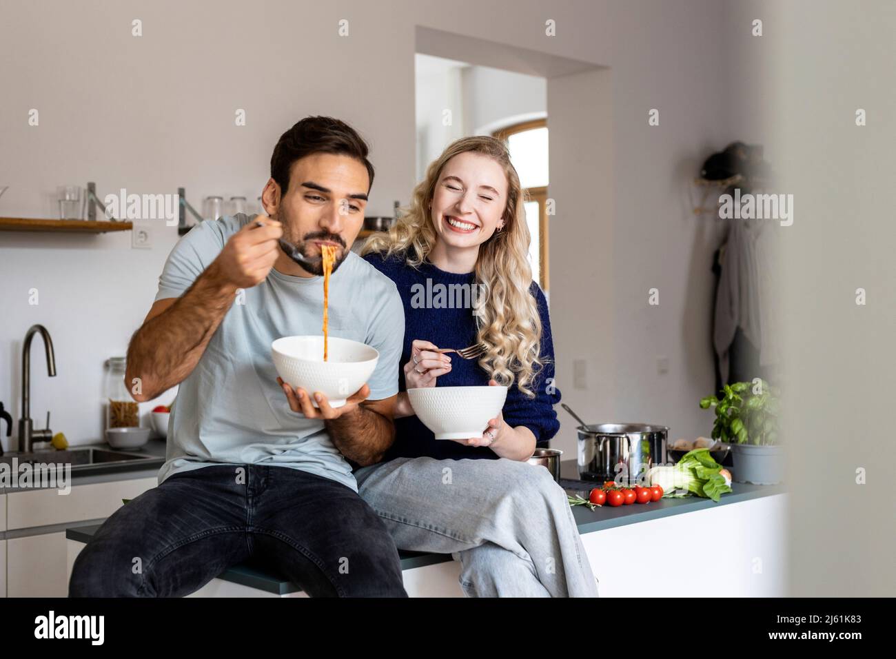 Smiling woman sitting by man eating noodles from bowl in kitchen at home Stock Photo
