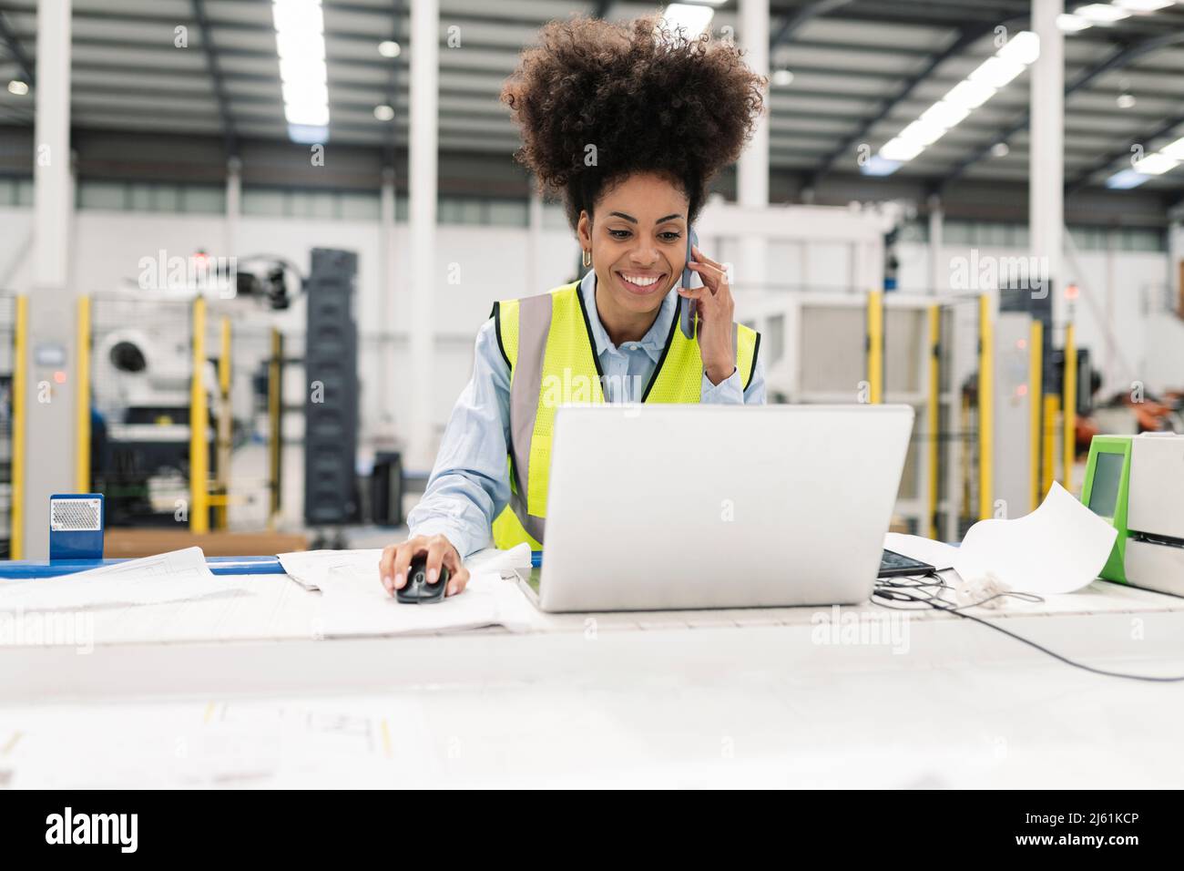 Smiling engineer working on laptop talking on mobile phone in factory Stock Photo