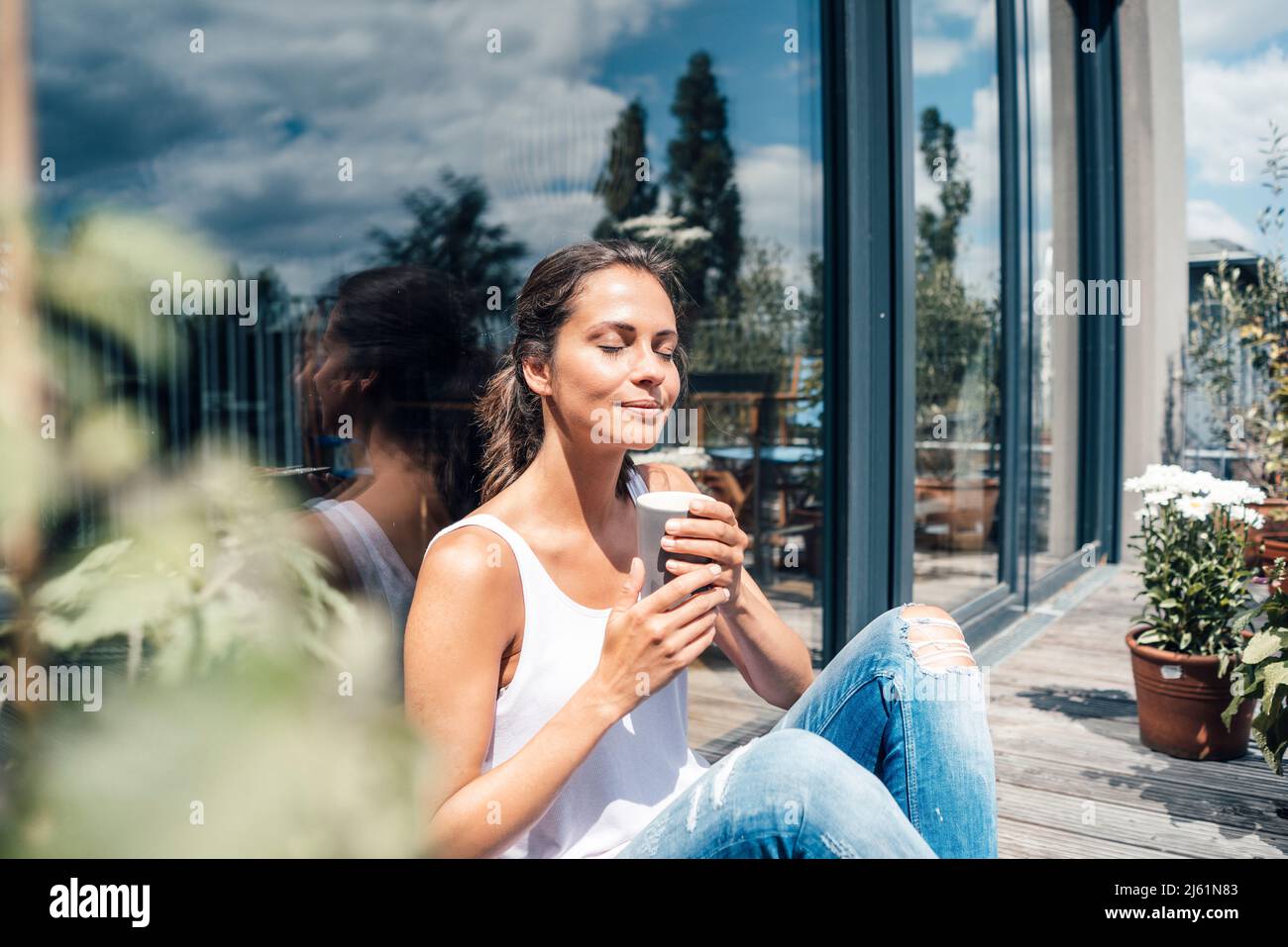 Smiling woman with eyes closed holding disposable coffee cup sitting in front of glass window at balcony Stock Photo