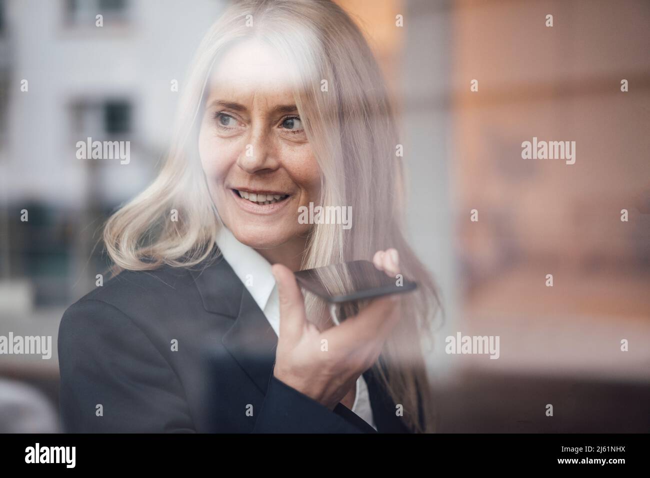 Businesswoman sending voicemail over smart phone seen through glass of work place Stock Photo