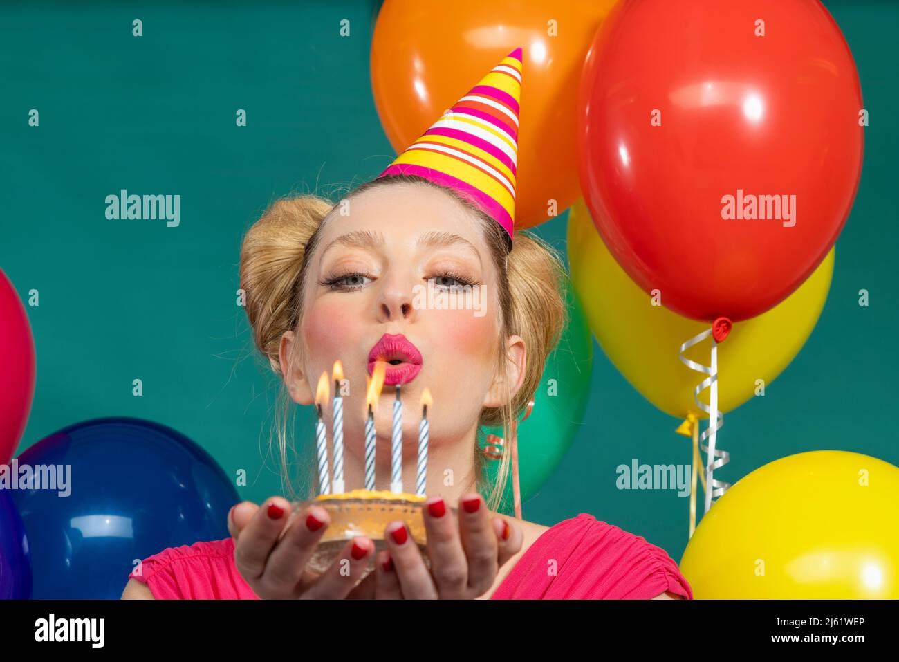 Beautiful young woman wearing party hat blowing candles on birthday cake against green background Stock Photo