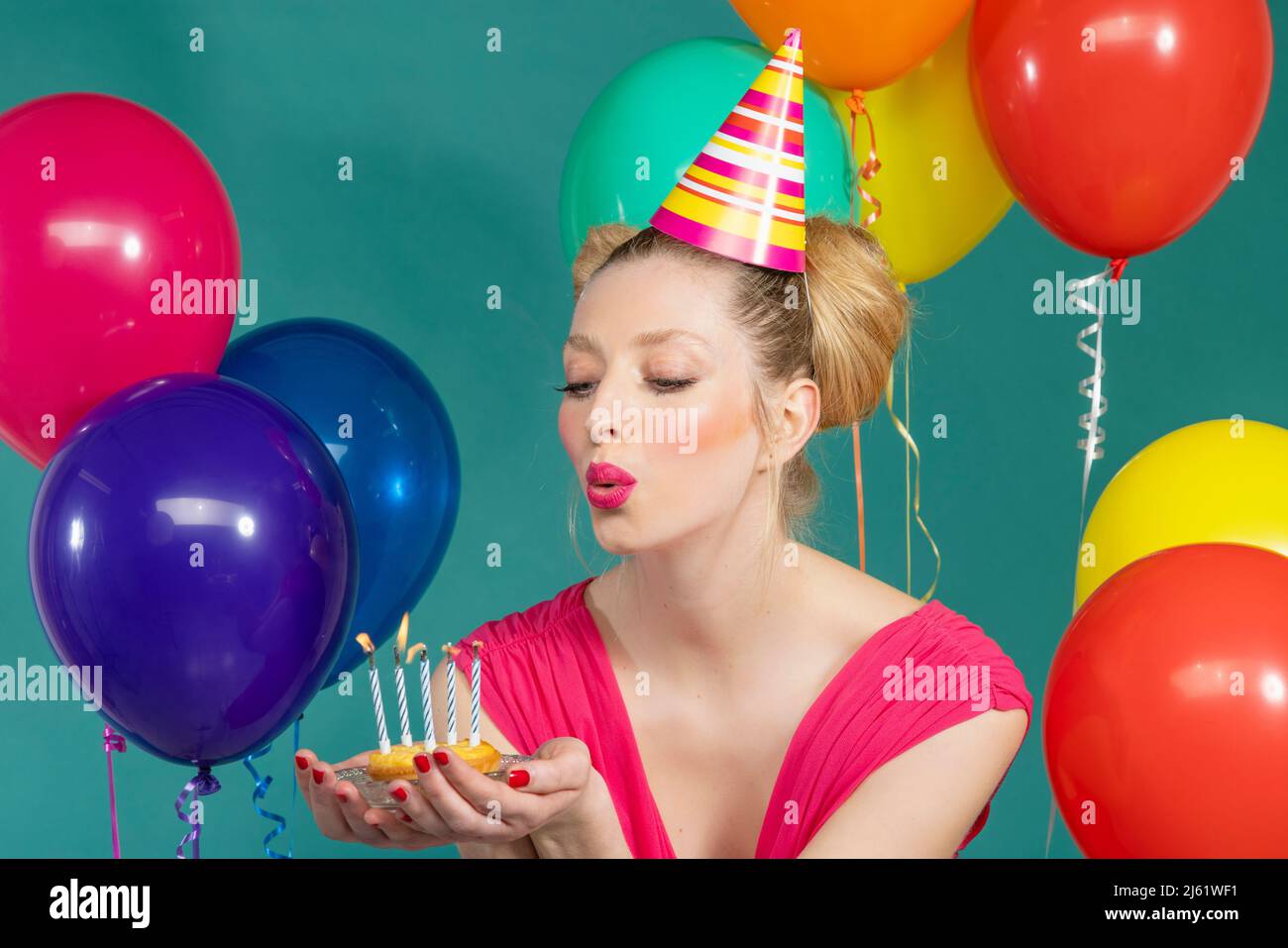 Young woman wearing party hat blowing candles on birthday cake against green background Stock Photo