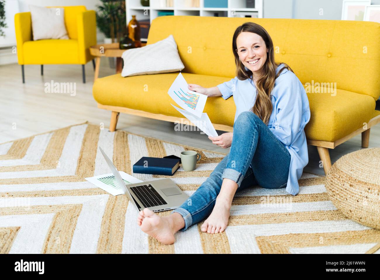 Happy freelancer with laptop and document sitting on carpet by sofa at