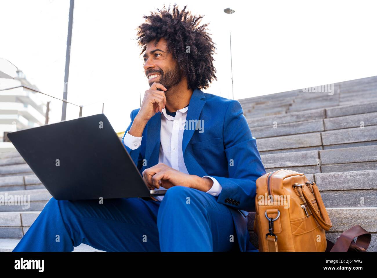 Smiling businessman holding laptop sitting with hand on chin by ...