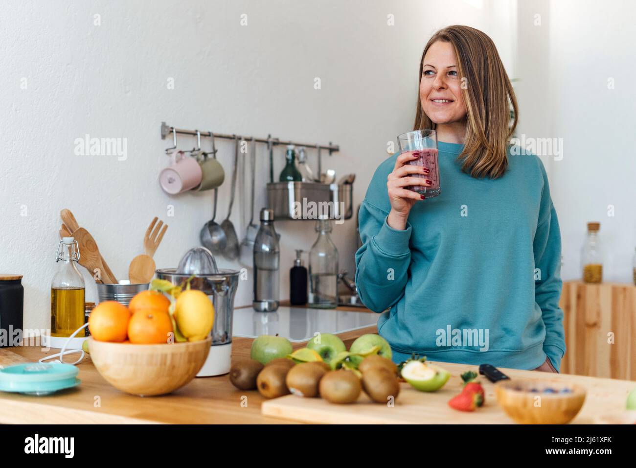 Smiling blond woman with glass of smoothie standing at table in kitchen Stock Photo