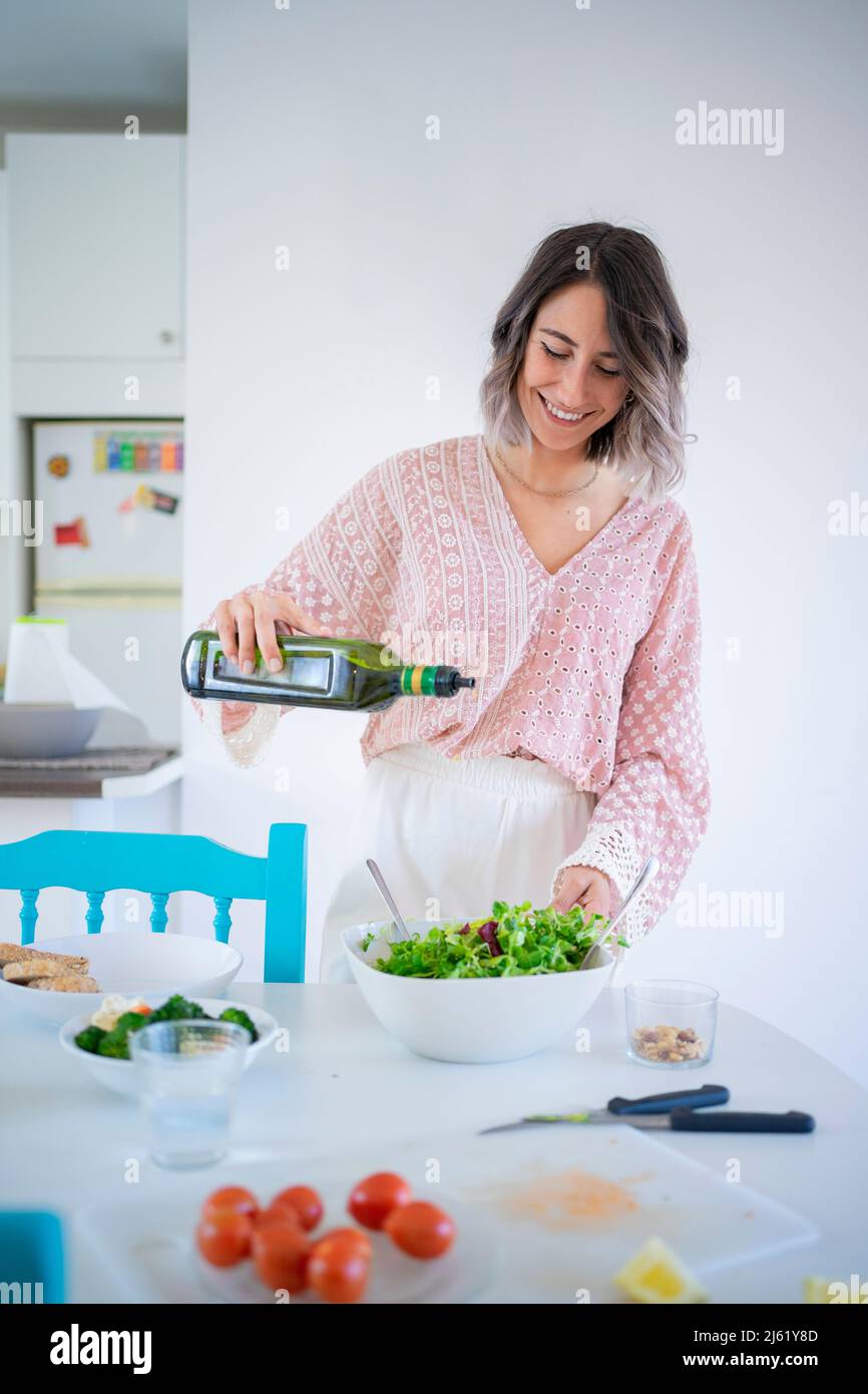 Smiling young woman pouring olive oil in salad bowl at table Stock Photo