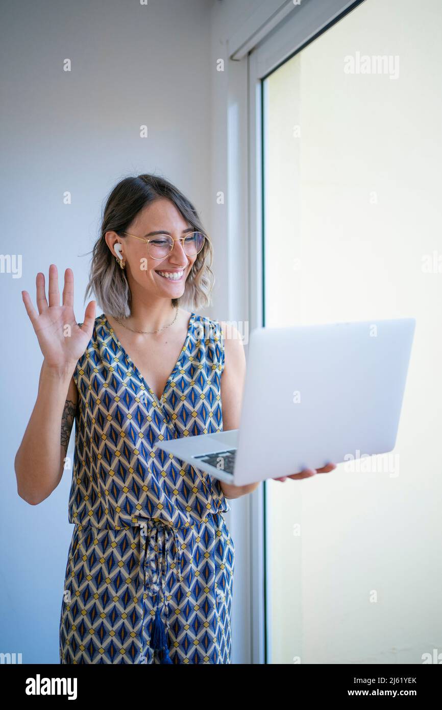 Happy freelancer waving hand on video call through laptop by window at ...