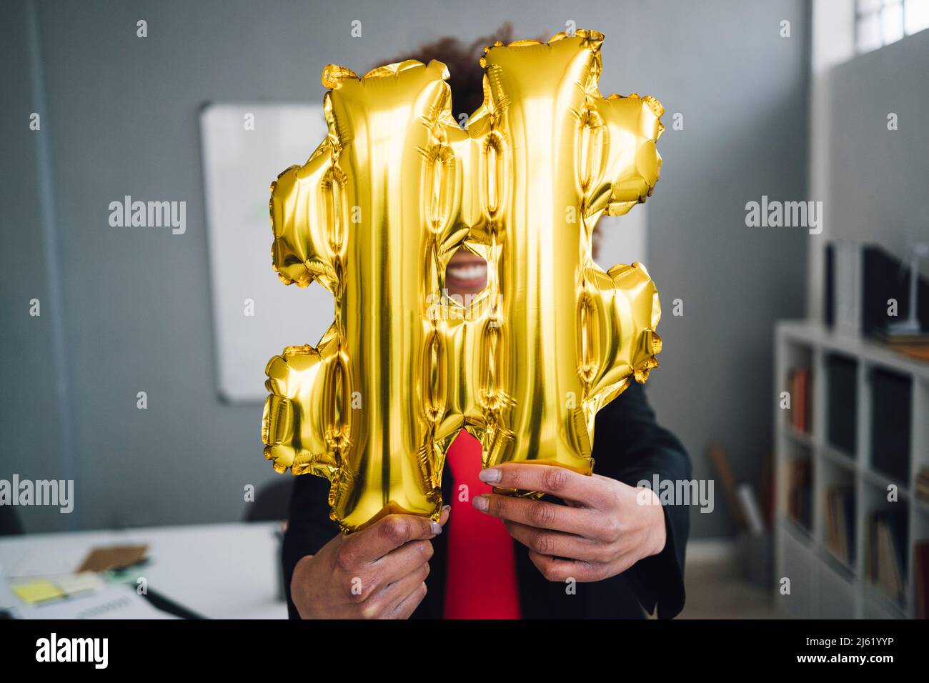 Businesswoman holding golden hashtag symbol balloon in front of face Stock Photo