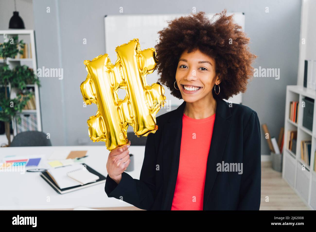 Smiling businesswoman with golden hashtag symbol balloon in office Stock Photo