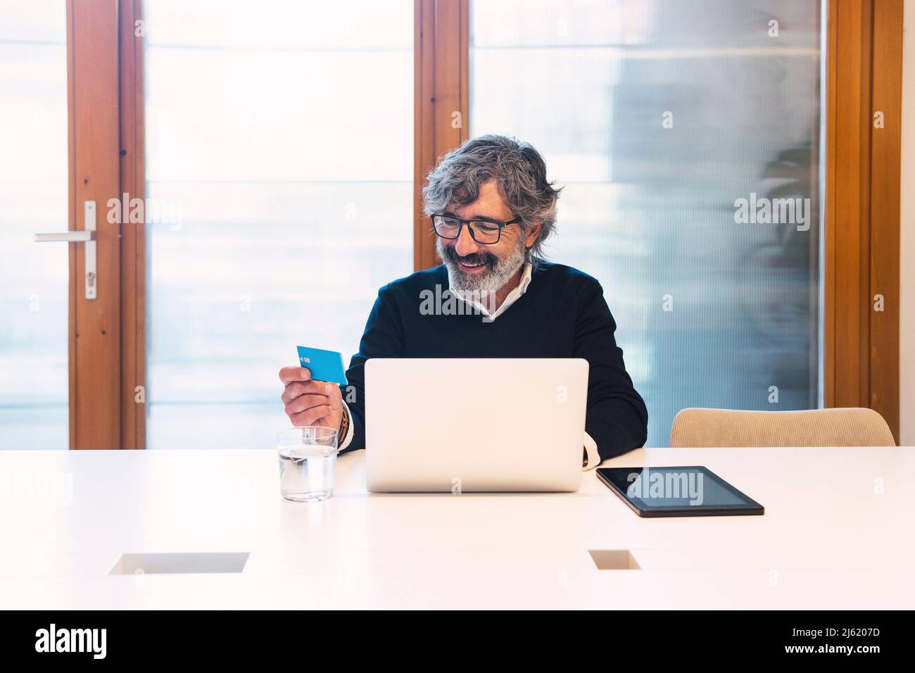 Happy senior freelancer wearing eyeglasses holding credit card sitting with laptop at table Stock Photo