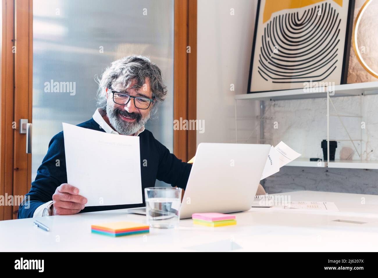 Smiling freelancer reading documents sitting with laptop at table Stock Photo