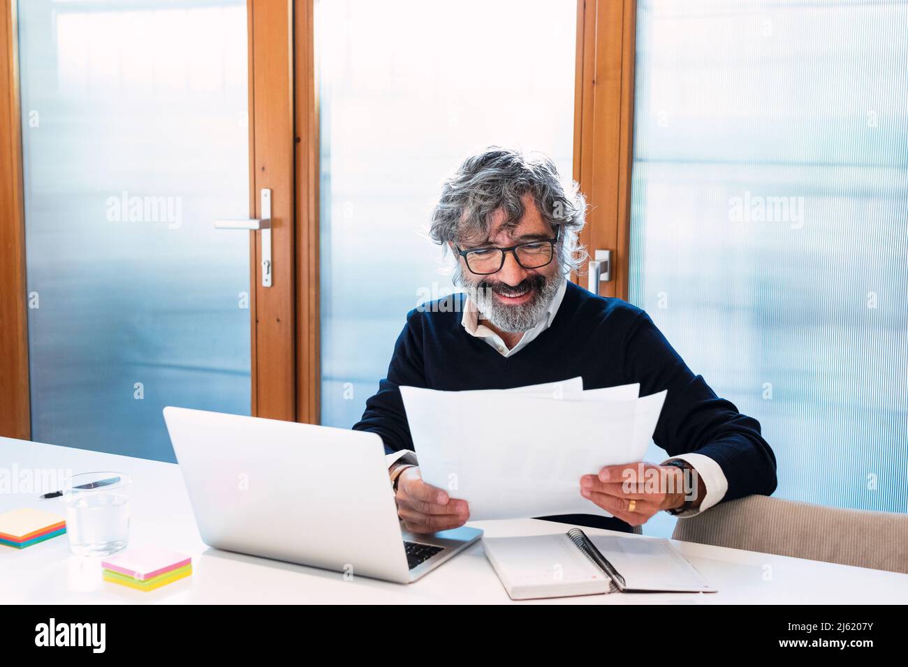 Happy freelancer reading documents sitting with laptop at home Stock Photo