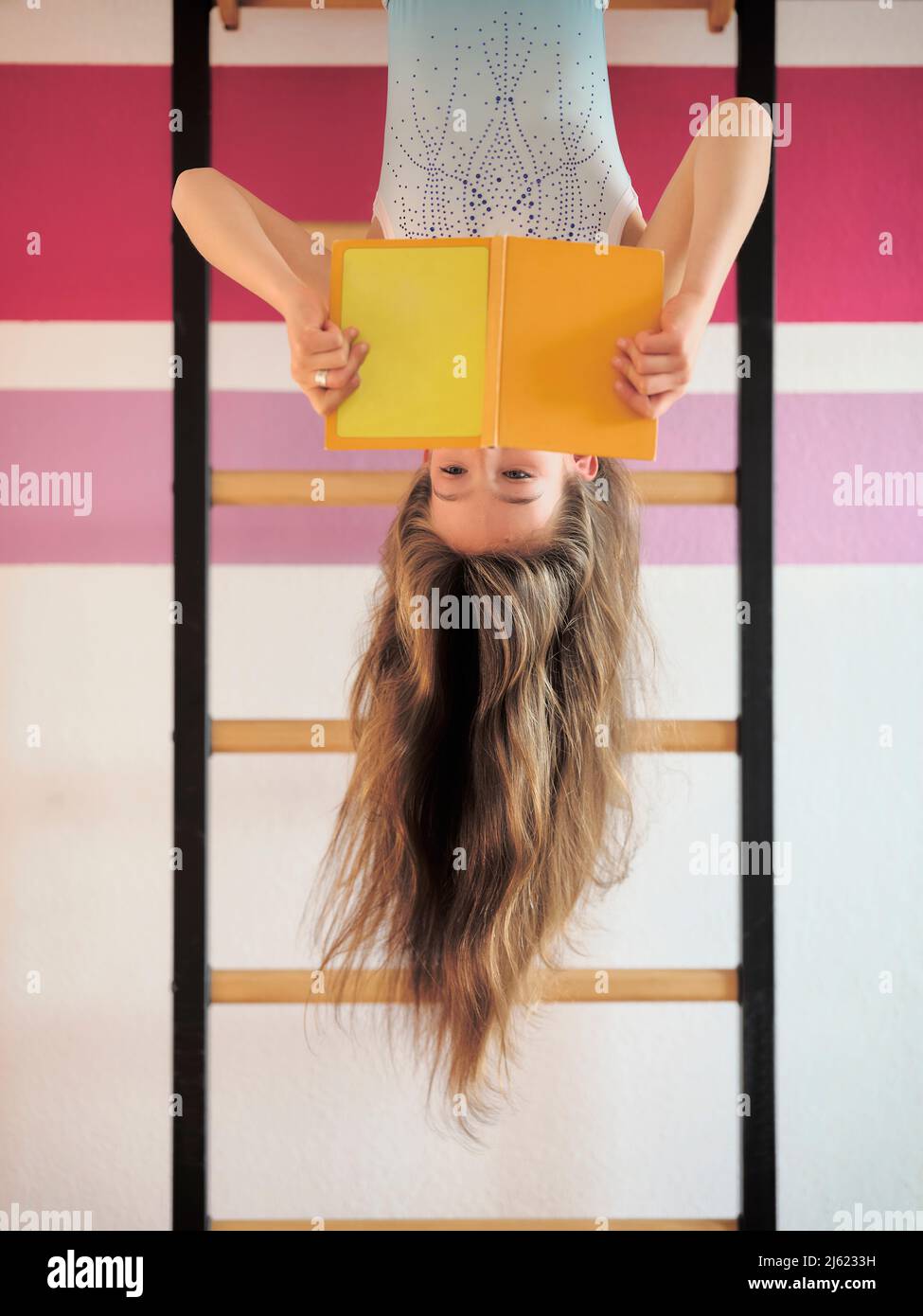 Girl reading book hanging upside down on wall bars Stock Photo