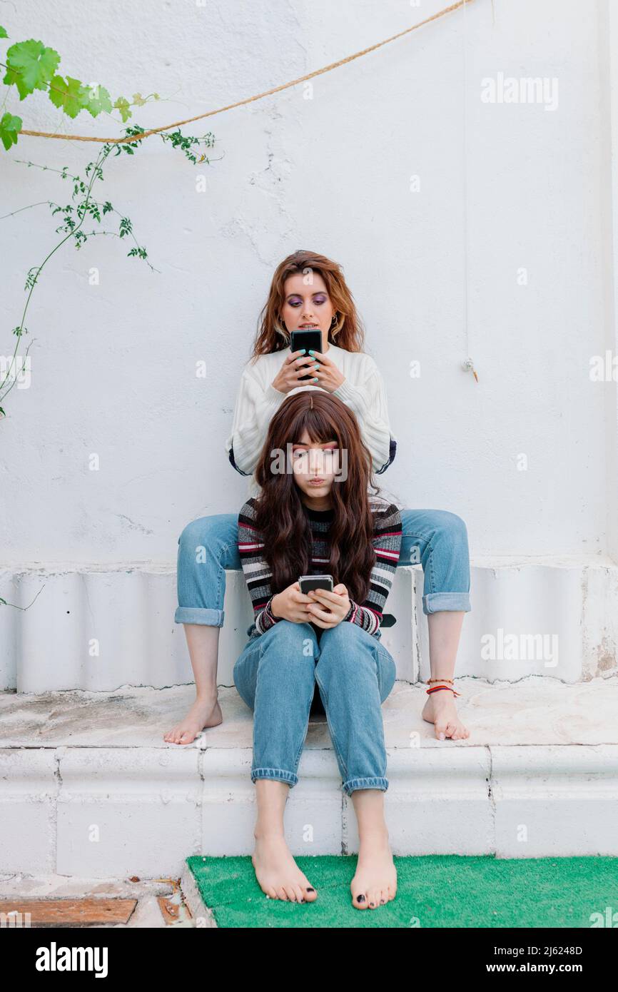 Friends with mobile phones sitting in front of white wall at back yard Stock Photo