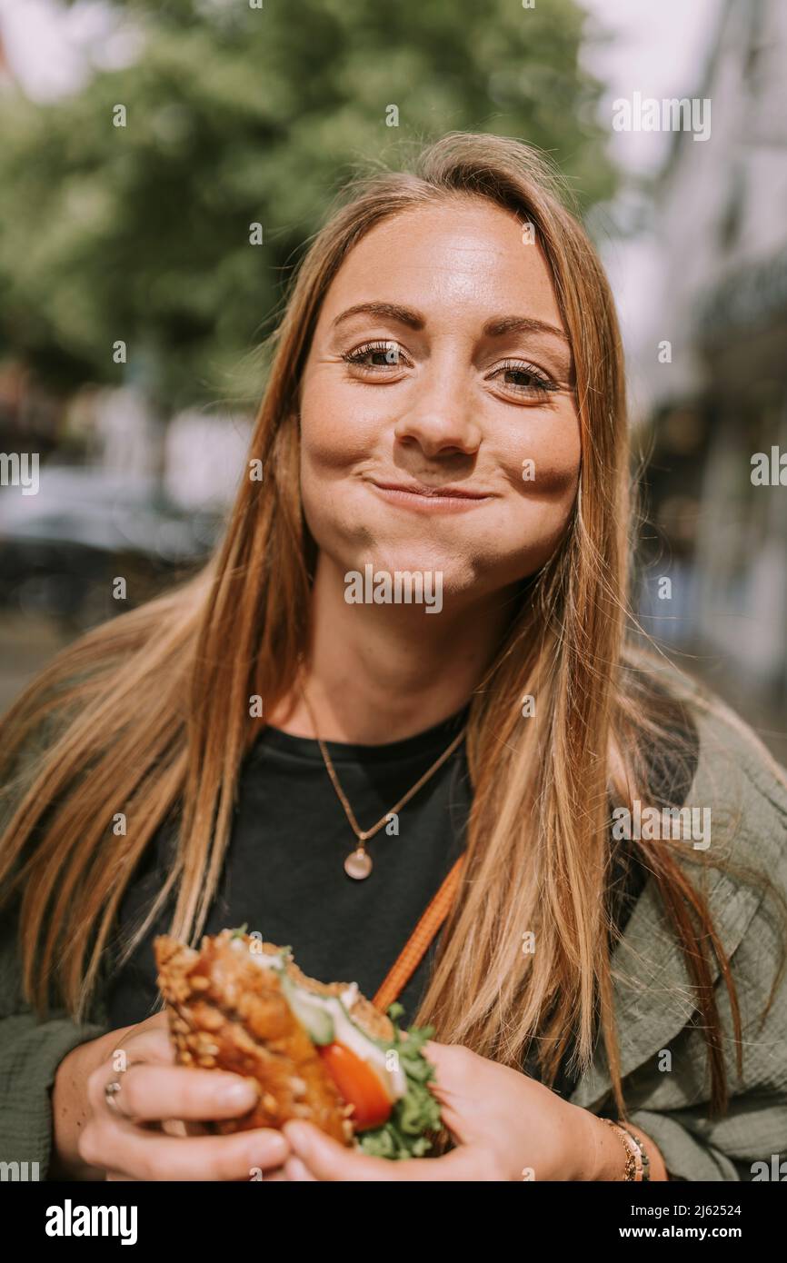 Smiling young woman with puffed cheeks eating sandwich Stock Photo