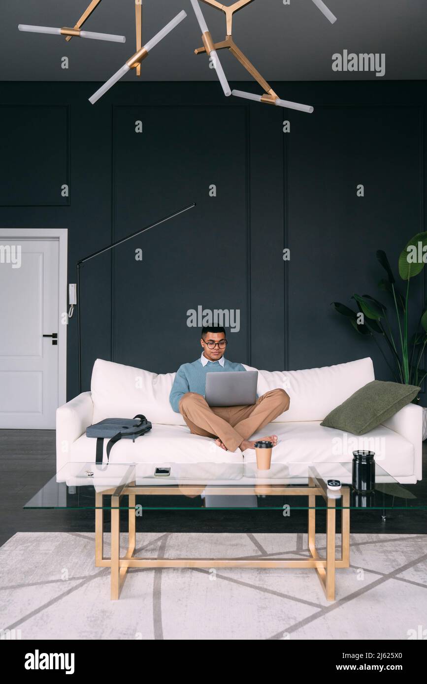 Freelancer working on laptop sitting cross-legged on sofa at home Stock ...