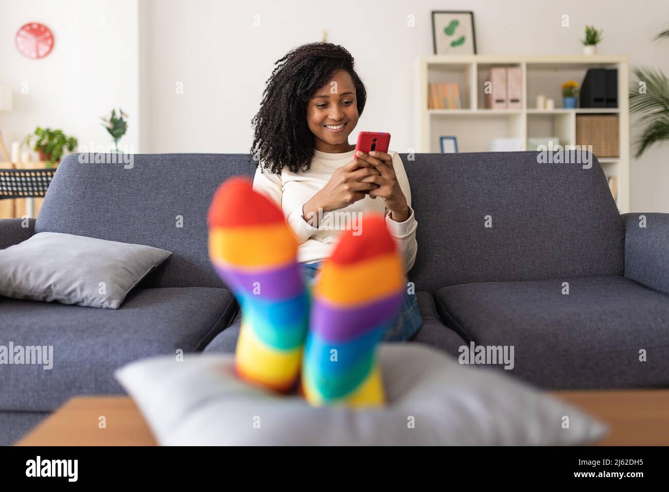 Smiling young woman using a mobile phone while relaxing on sofa at home. Stock Photo