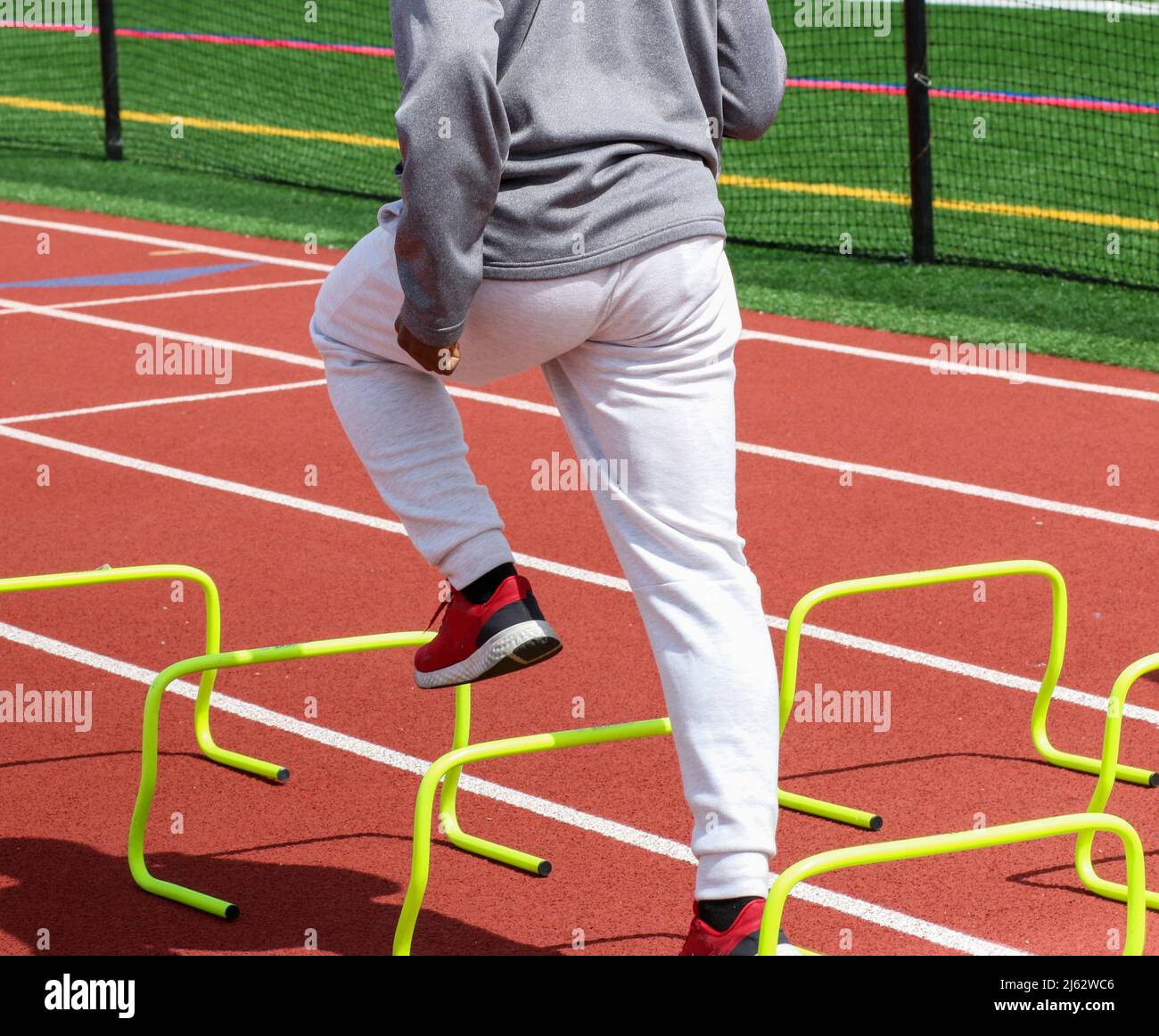 Rear view of a high school boy standing over yellow hurdles on a red