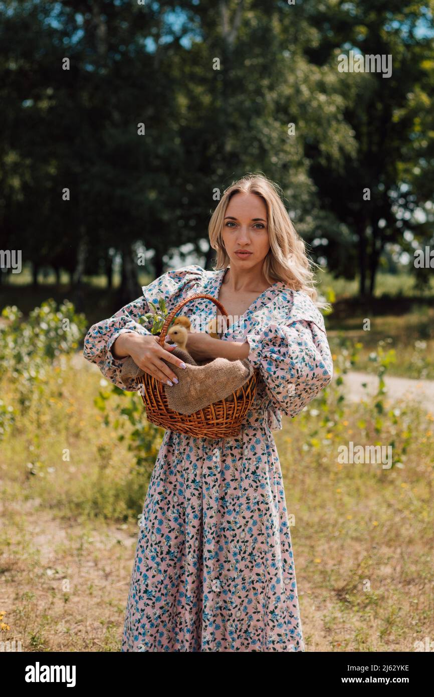 A young woman in long dress holds a basket of ducklings and smiles Stock Photo