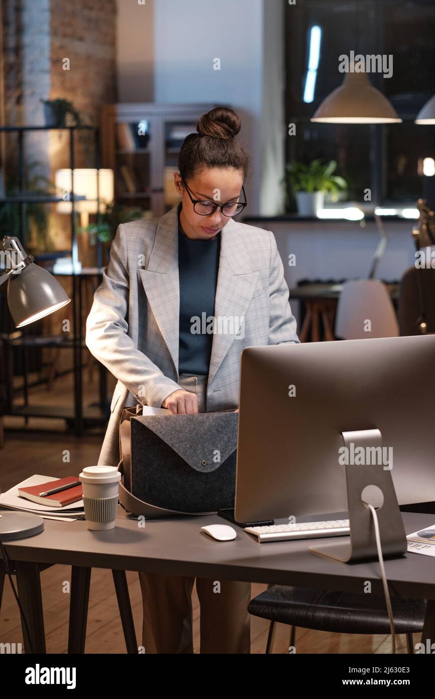 Serious young mixed race manager in glasses standing at table with computer and taking work staff while leaving office at night Stock Photo