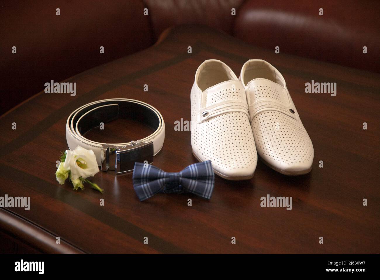 Groom's white shoes, belt, bow tie and boutonniere, top view. Groom's morning, wedding details, preparation for the wedding ceremony. Stock Photo