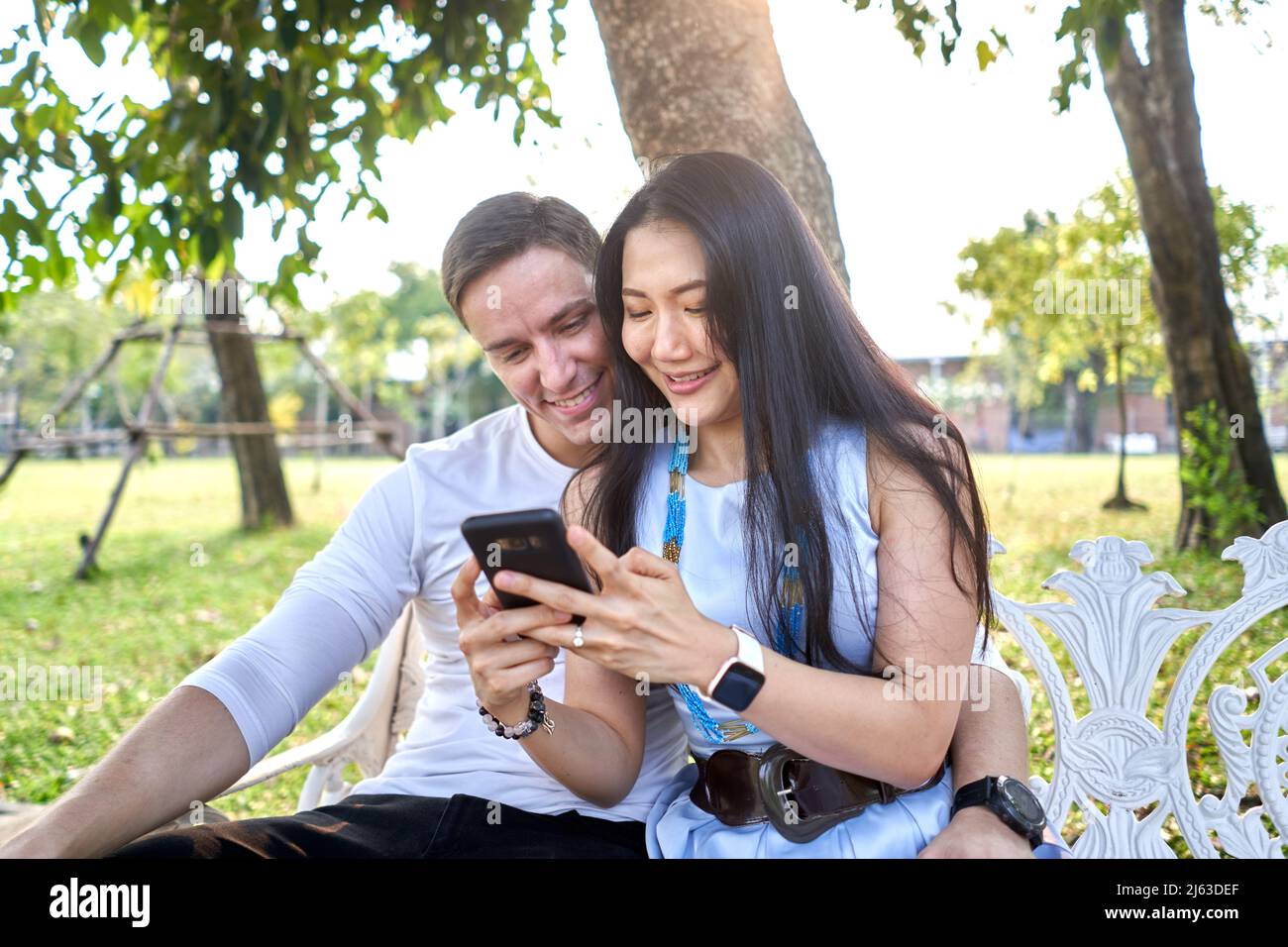 Smiling couple using the mobile while sitting on a bench outdoors during sunset Stock Photo