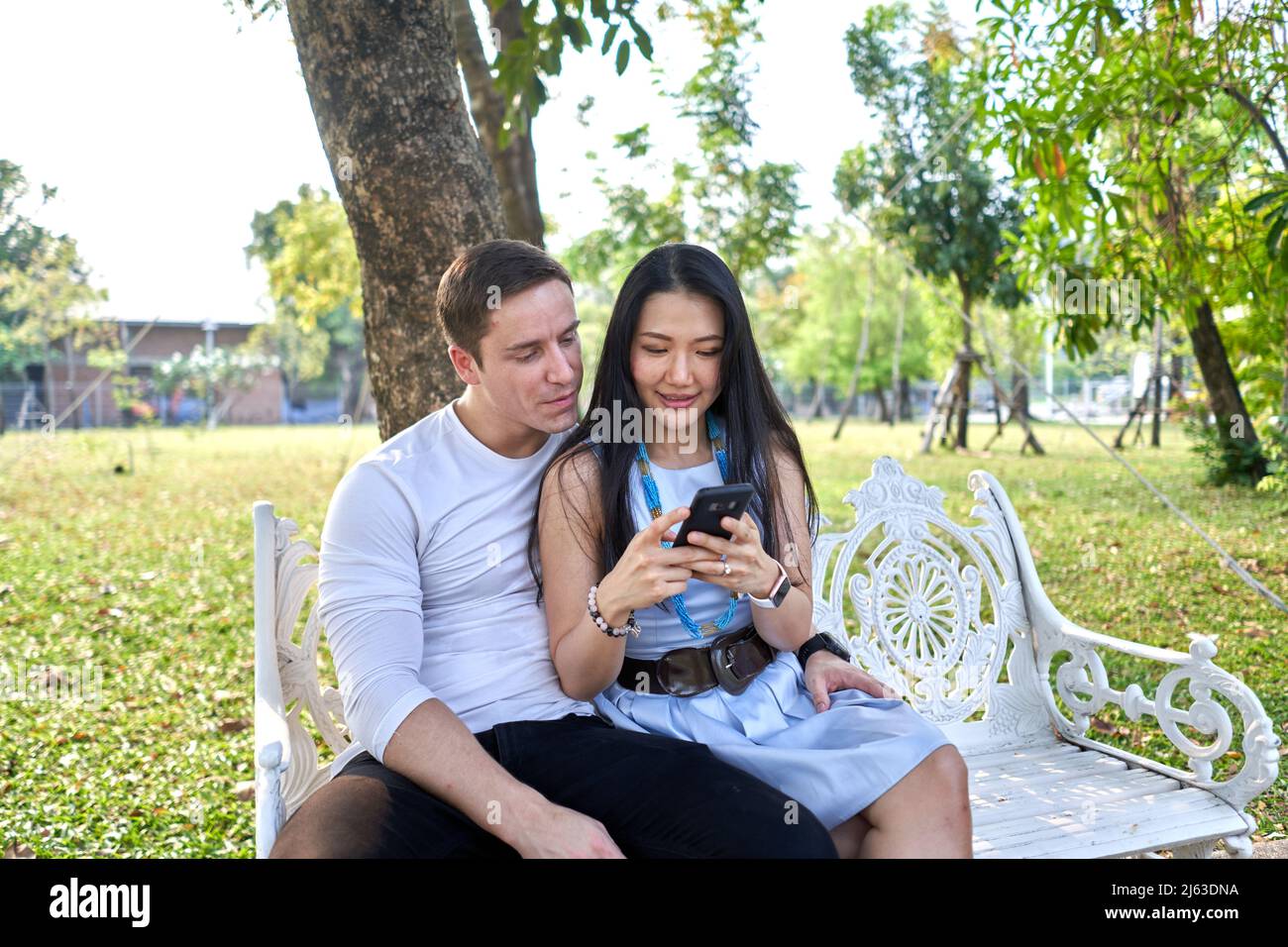 Couple sitting on a white bench in a park using a mobile during sunset ...