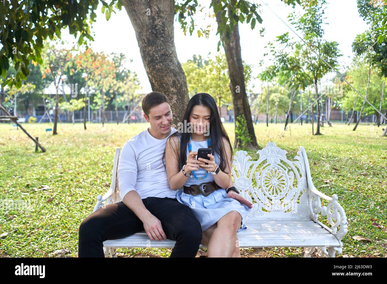 Multiethnic couple sitting together on a bench while using the mobile outdoors Stock Photo