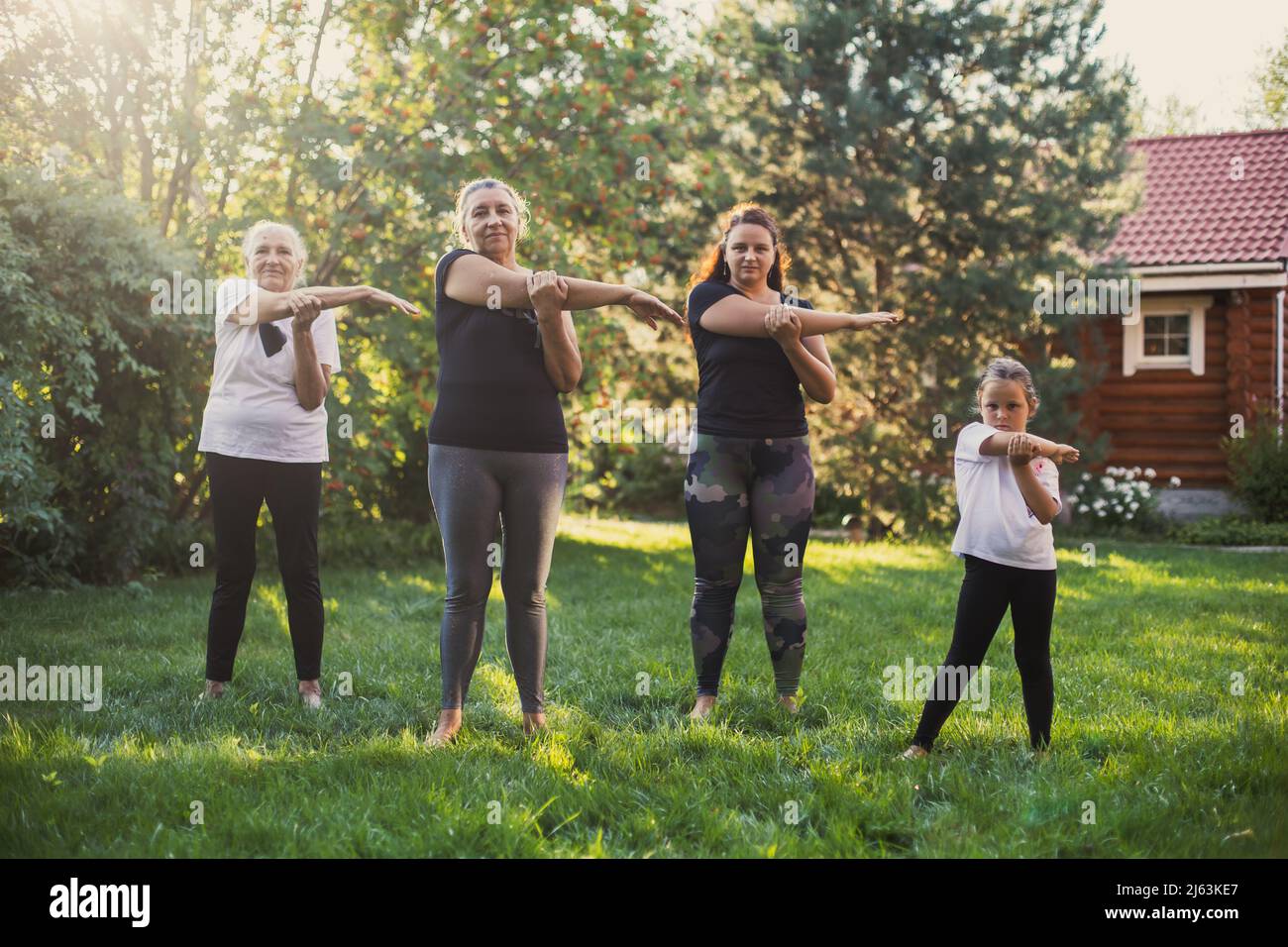 Females of few generations exercising outside stretching arms together in same way standing on meadow full of green grass and trees. Spending time Stock Photo