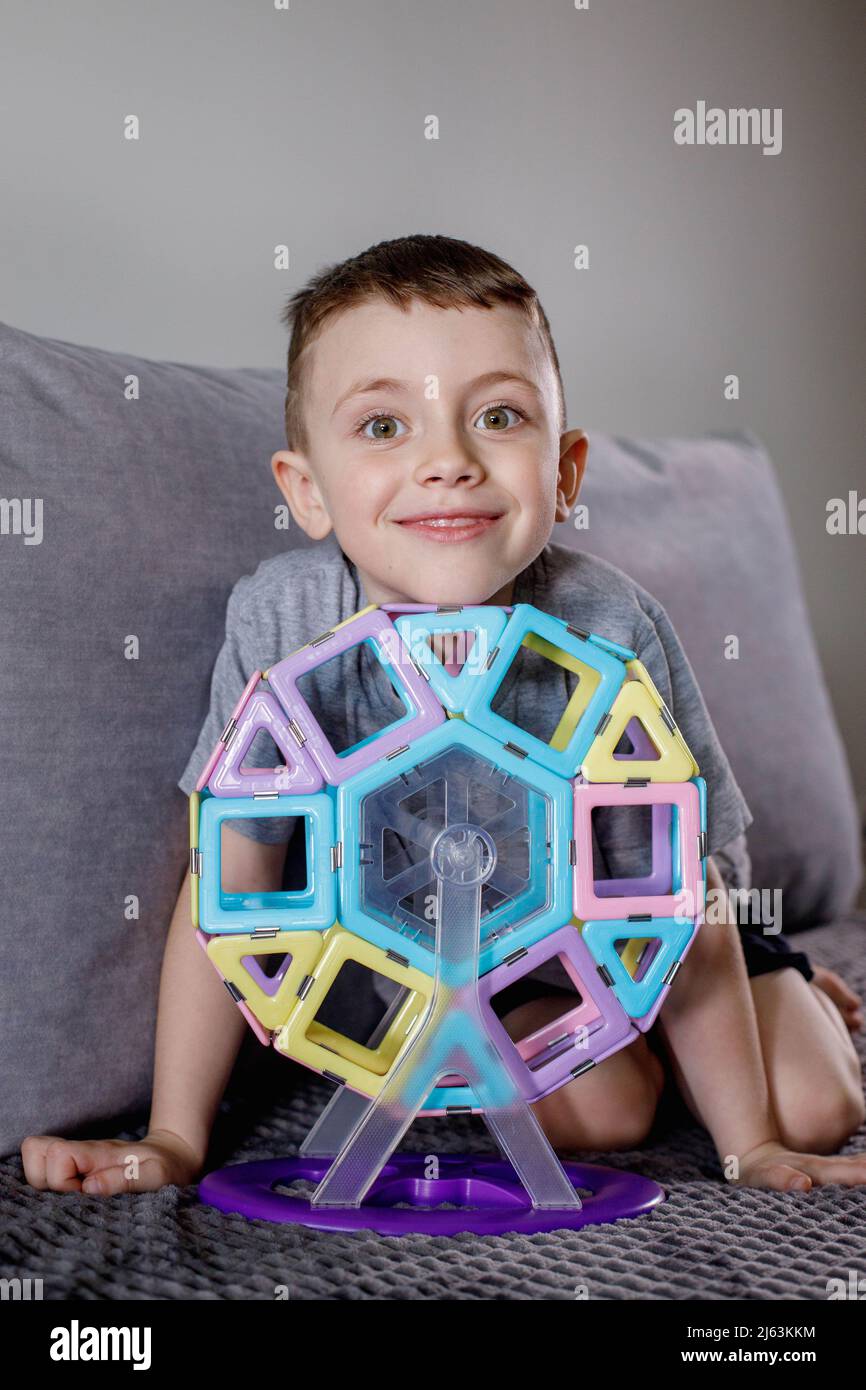 A boy in a gray Tshirt sits on a bed near a white wall and plays with a Ferris wheel, which he
