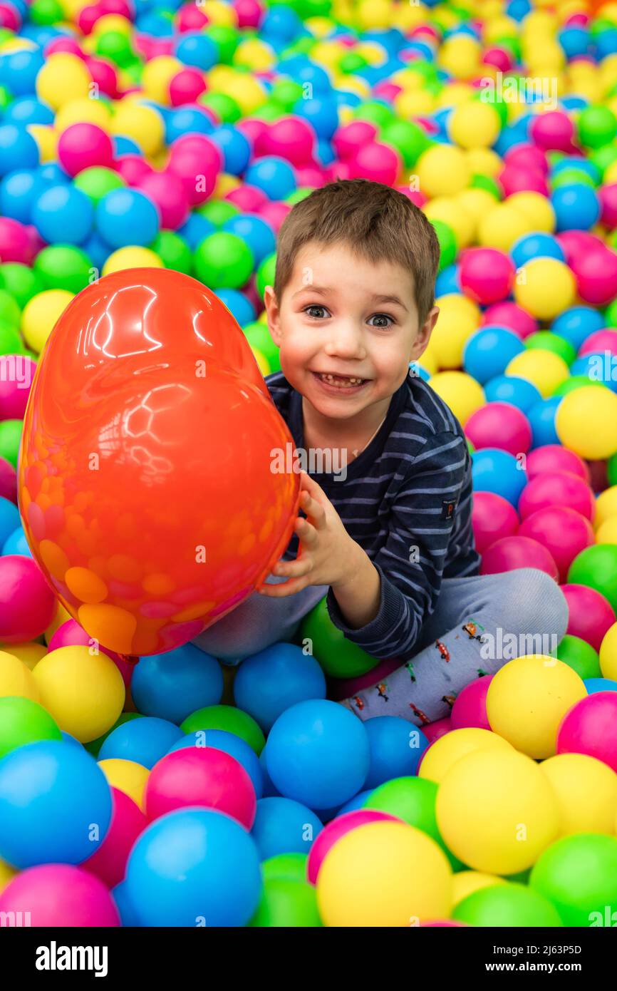 A little boy sits in bright balls and holds in his hands a red ball