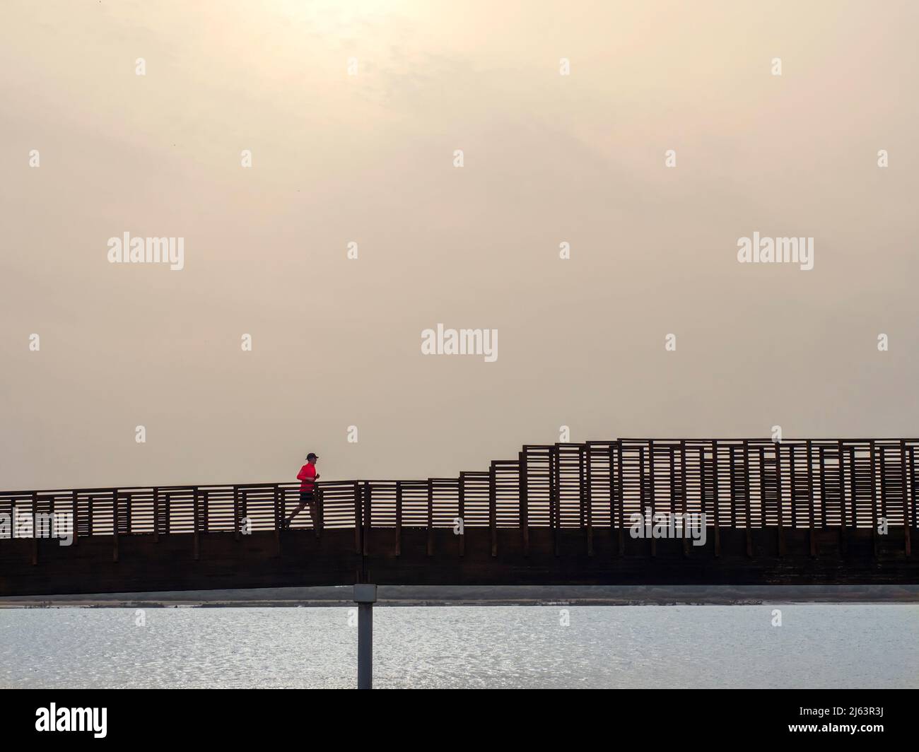 Man in red t-shirt running along the raised wooden boardwalk of the Odiel river in Huelva, on a sunny day Stock Photo