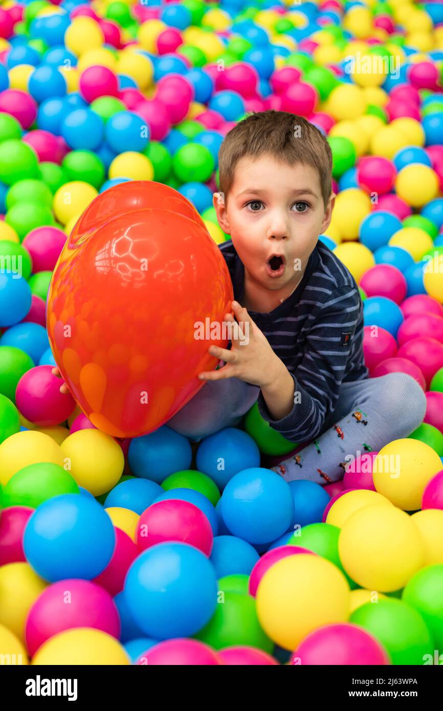 A little boy sits in bright balls and holds in his hands a red ball