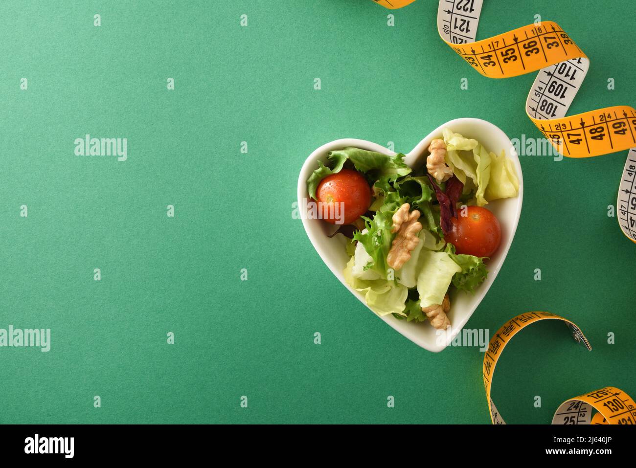 Concept of diet and health with heart-shaped bowl with salad with nuts and metric tape on a green background. Top view. Horizontal composition. Stock Photo
