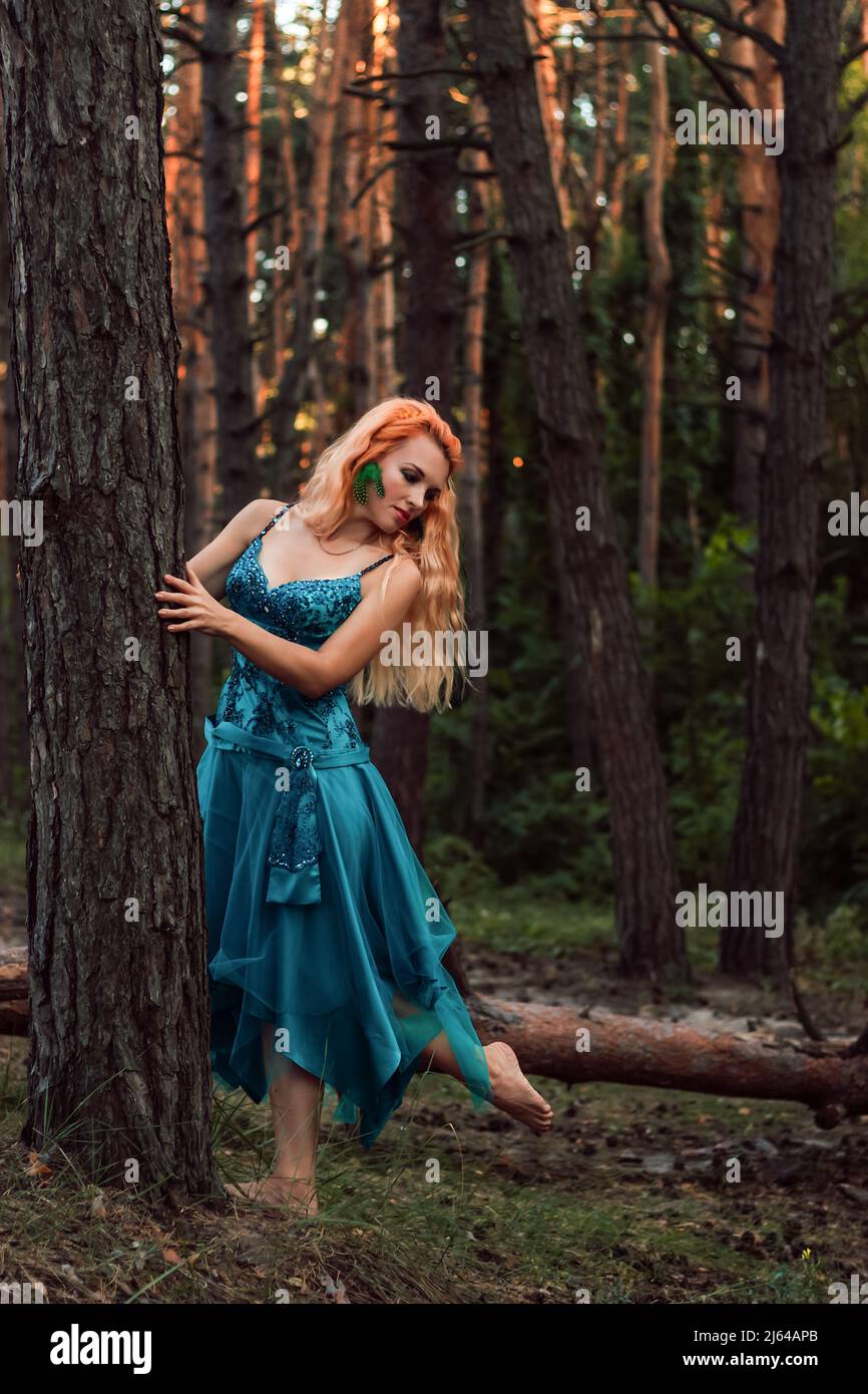 A beautiful girl with red hair stands near a tree in the forest and ...