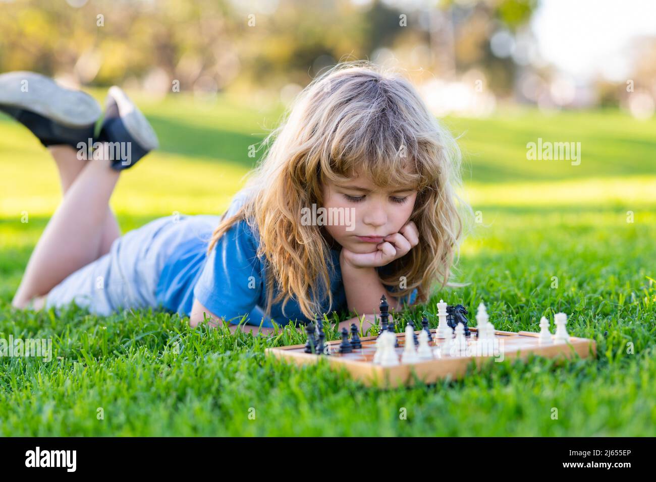 Clever concentrated and thinking child while playing chess, laying on grass in summer park. Chess, success and winning. Stock Photo