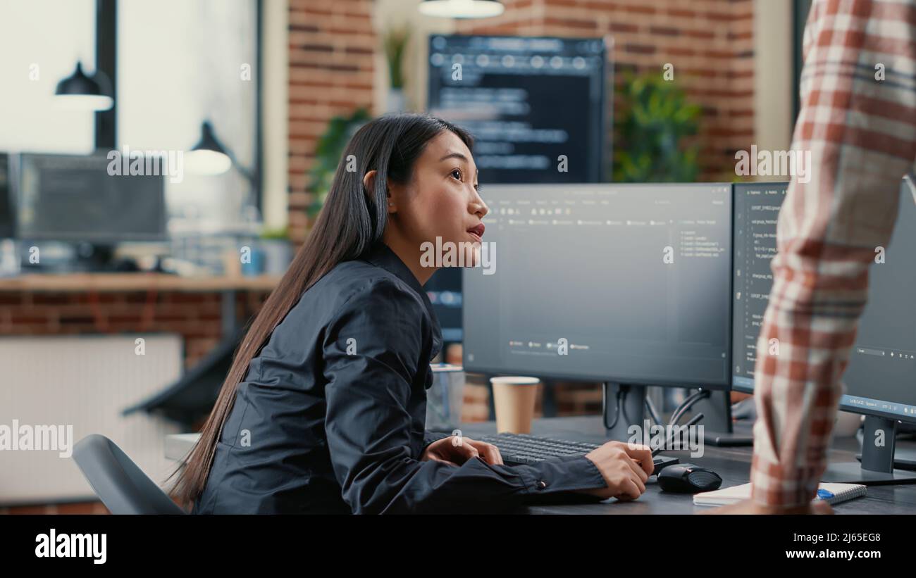 Database developer pointing pencil at computer screen with software compiling code explaining source algorithm to coworker standing next to desk. System engineers discussing ai team project. Stock Photo