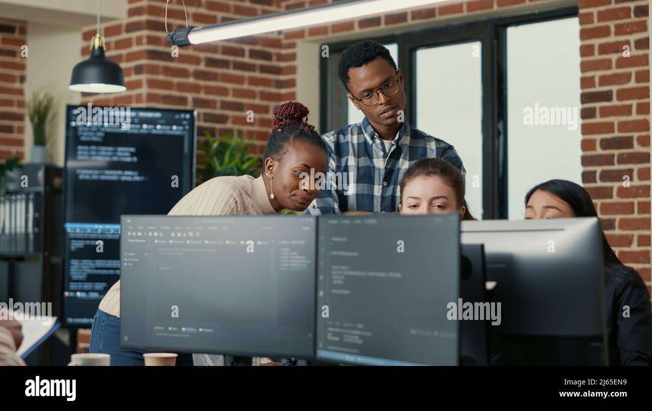 Developers brainstorming ideas looking at code on computer screens asking feedback from senior developer while intern joins the discussion. Junior programmers collaborating on group project. Stock Photo