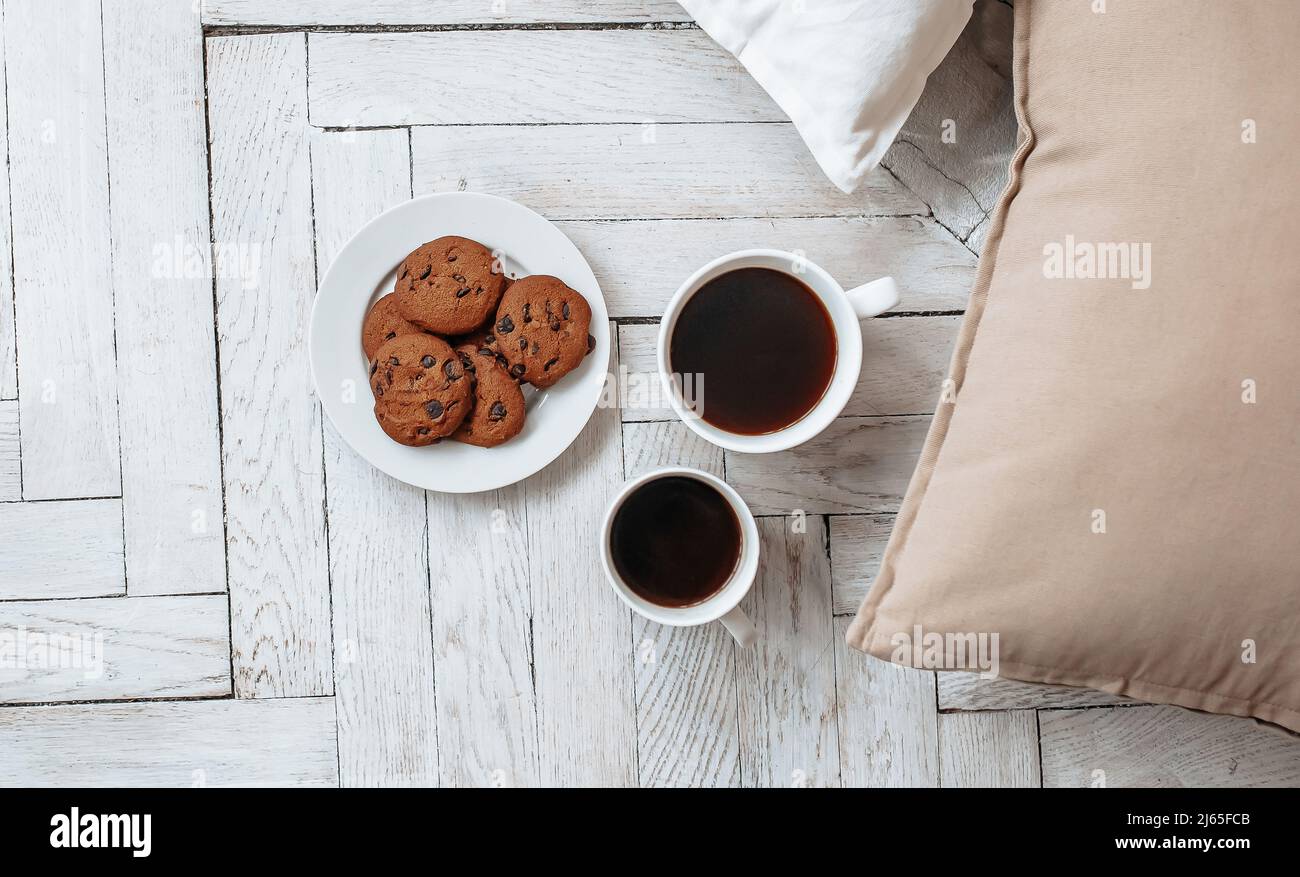 Breakfast in the morning. Two white cups of coffee and chocolate chip cookies stand on a light wooden parquet and pillows lie nearby. Homeliness, day Stock Photo
