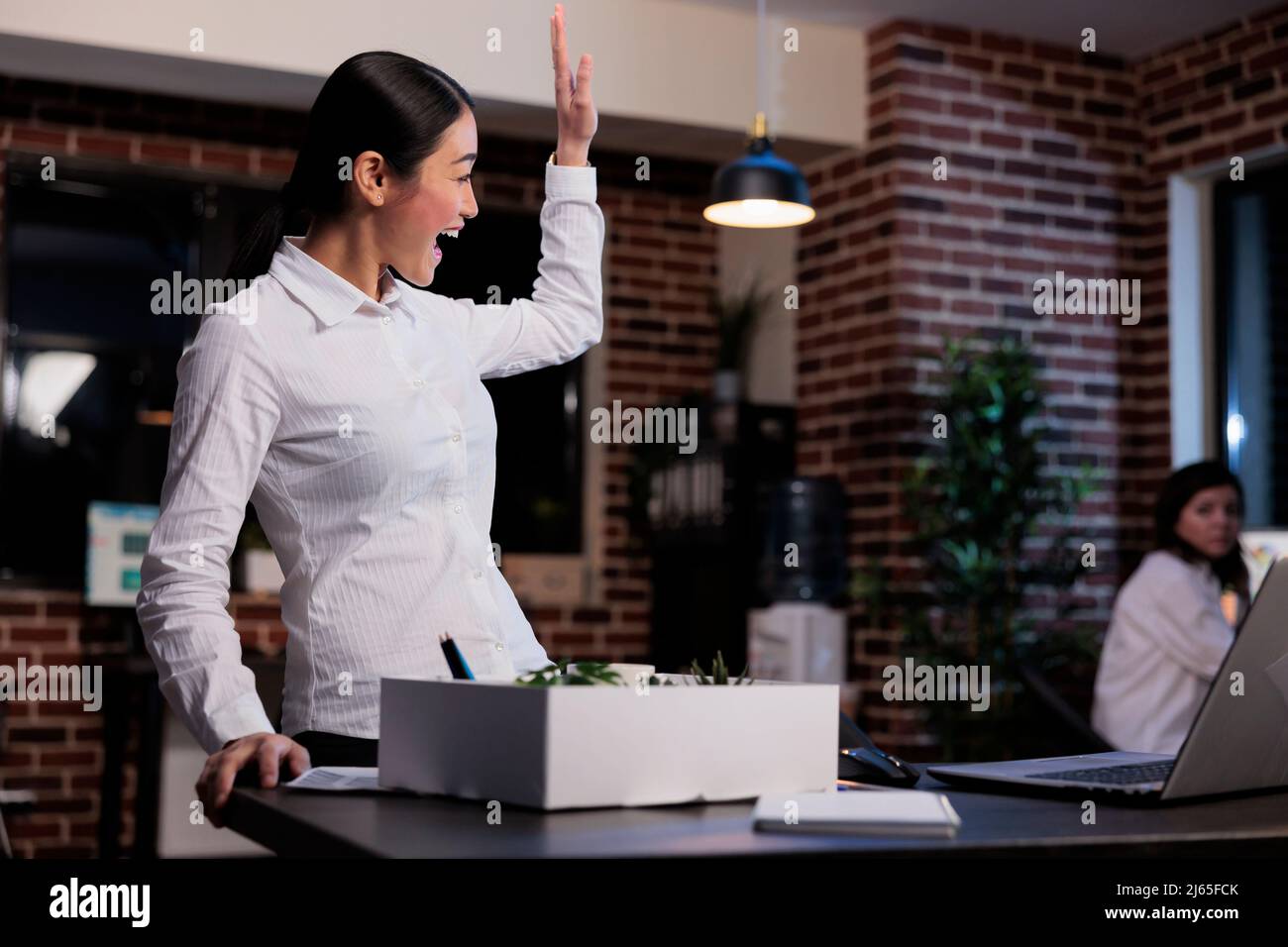 Fired businesswoman packing personal belongings while waving goodbye to office colleague. Dismissed business company employee putting personal stuff in cardboard box because of project failure. Stock Photo