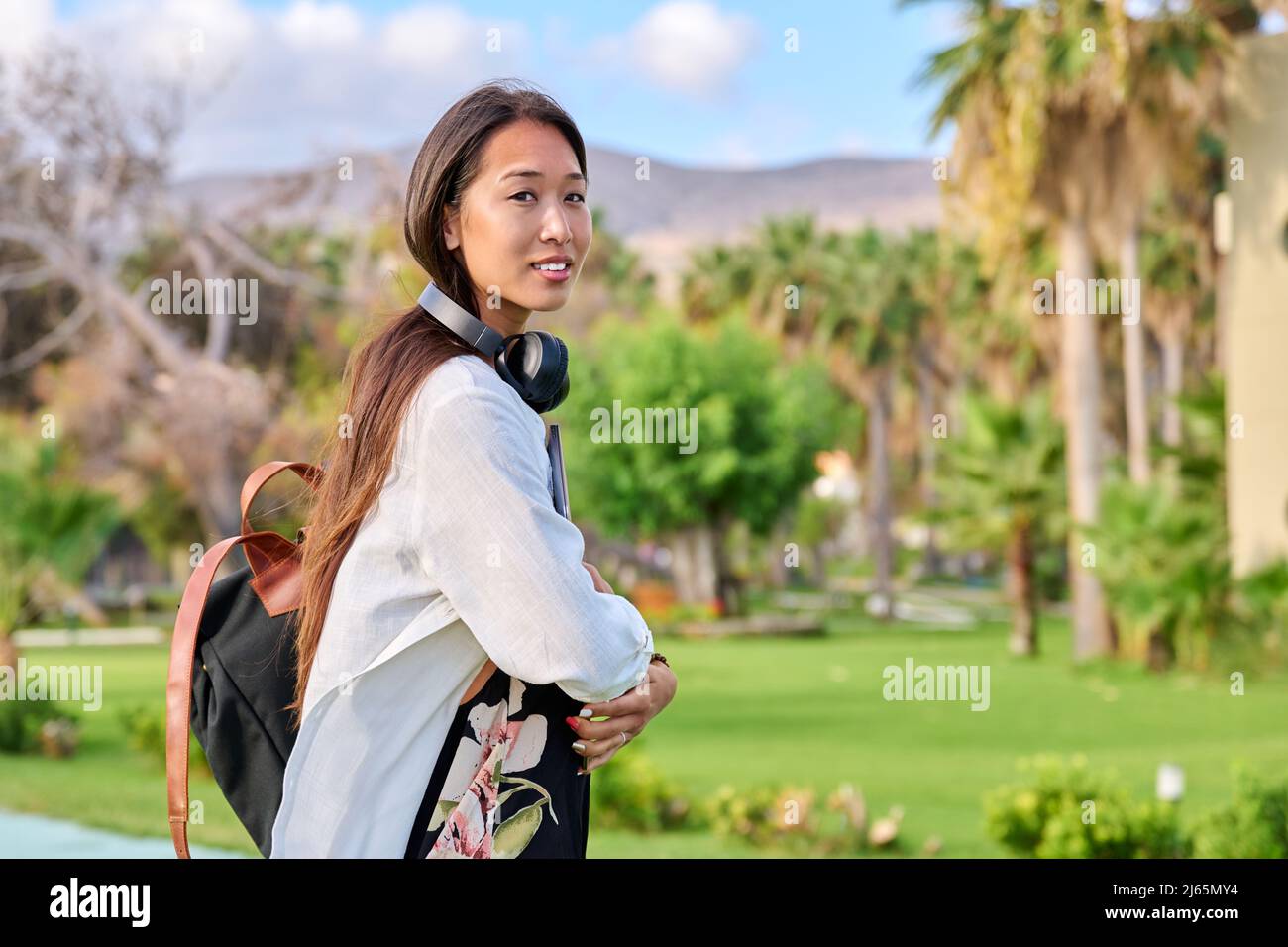 Outdoor portrait of asian female student with backpack headphones laptop looking at camera Stock Photo