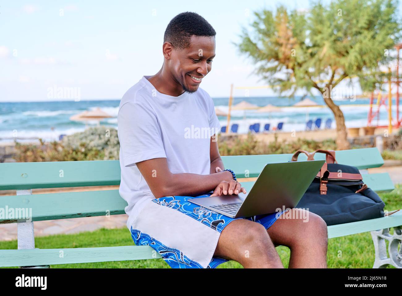 Young smiling male freelancer relaxing on beach, sitting on bench using laptop Stock Photo
