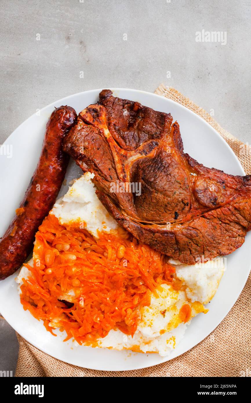 Flat lay of Traditional South African Braai or Shisa nyama, meat cooked on an open flame and traditional side dish of pap and chakalaka. Stock Photo