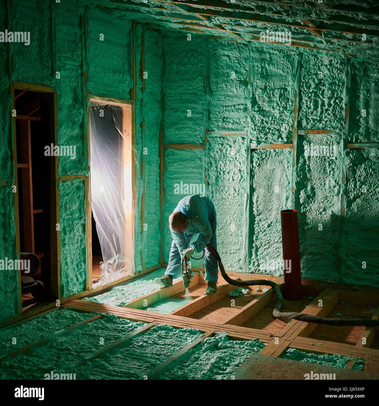 Male builder insulating wooden frame house. Man worker spraying polyurethane foam on floor inside of future cottage, using plural component gun. Construction and insulation concept. Stock Photo