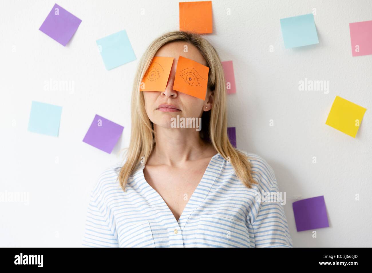 Business woman sits in bright modern office behind her desk on which are many office things and has sticky notes with drawn eyes on her face and sleep Stock Photo